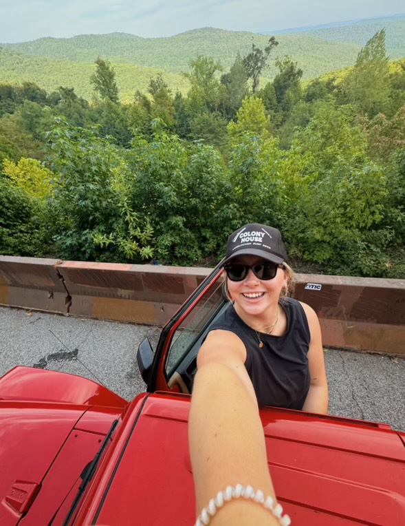 Everett takes a selfie in a black hat outside of her red Jeep with a forest in the background