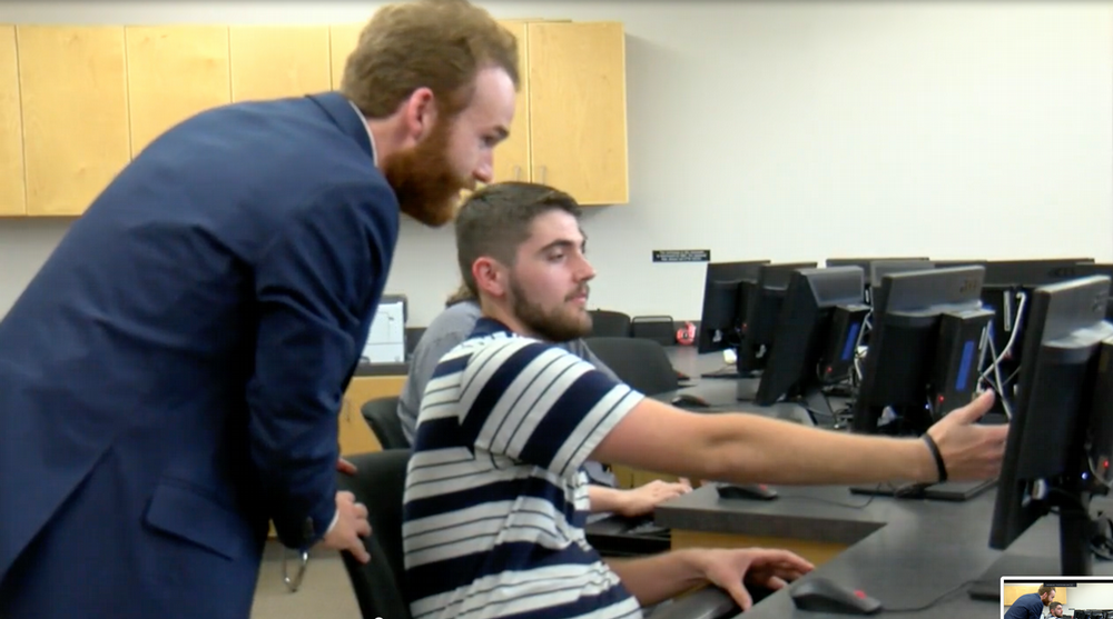 Joe Faith stands behind student Landen Wilson to help with his work on a computer in a campus lab.