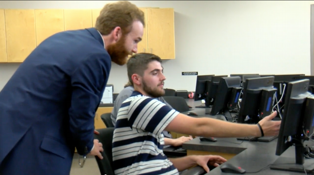 Joe Faith stands behind student Landen Wilson and helps him with work on his computer screen in a campus lab.