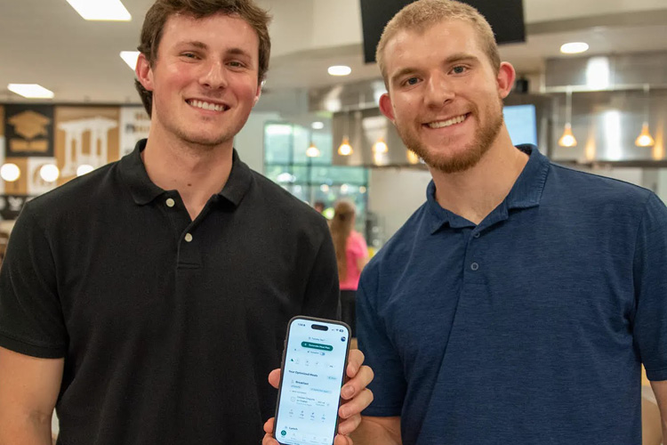 Students Simon Bertolino and Fletcher Stobaugh stand in the cafeteria showing their AI developed app on a smart phone.